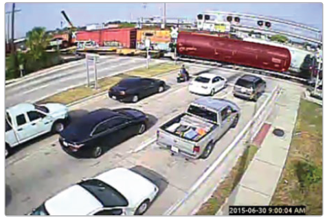 Vehicles stopped at a highway-railway grade crsossing waiting for a series of tanker cars to pass.