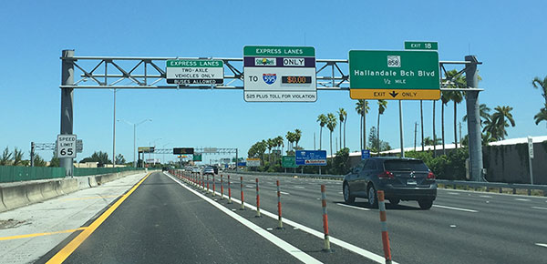 Photograph of the 95 Express in Miami, Florida.  The photo shows the northbound lanes of the I-95 corridor from the vantage point of the 95 Express.  A set of orange plastic delineators separate the 95 Express from the general purpose lanes.  An overhead sign bridge shows a sign that states that two-axle vehicles and buses are permitted to travel in the 95 Express.  A second overhead signs shows text that states the 95 Express only permits travel from vehicles equipped with a SunPass toll transponder.  The second sign also shows a toll rate of $0.00 in a changeable text panel and '$25 Plus Toll for Violation' as a line of fixed text.