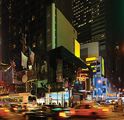 Photo. Nighttime street view of traffic driving through a brightly lit city intersection.