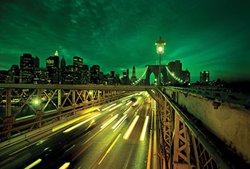 Photo. Nighttime view of traffic moving across a large bridge, heading into a downtown area. 