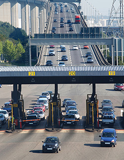 Photo. Front view of vehicles descending from a bridge and going through a toll collection station.