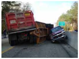 Photo of a major incident in which a jacknifed trailer has crushed a pickup truck against a line of concrete barriers, blocking the roadway.