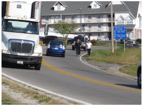 A truck and car pass each other moving in opposite directions on a two-lane curving road. The car must swing out over the double yellow line to pass two pedestrians walking on at the edge of the roadway.