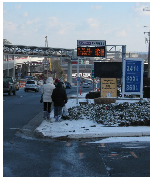 Pedestrians walking on a narrow, snow-covered sidewalk between a gas station from a major arterial. The landscaping of the gas station has grown out over the sidewalk, narrowing it further.