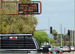 A busy arterial road with a travel time information sign mounted on an overhead gantry above the roadway indicating travel time to a significant point on the route.