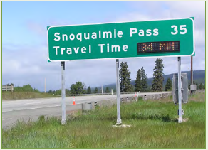 A busy arterial road with a travel time information sign mounted on an overhead gantry above the roadway indicating travel time to a significant point on the route.