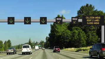 Picture (a mock up) of dynamic lane assignment, with dynamic message signs over each lane displaying green arrows or yellow diagonal arrows instructing traffic to merge out of those lanes.