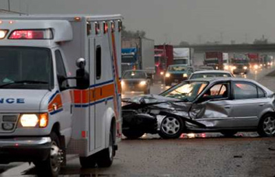 A photo of a car accident scene at the side of the highway. An emergency vehicle sits in front of a heavily damaged passenger car. Highway traffic is seen backed up behind the accident. 