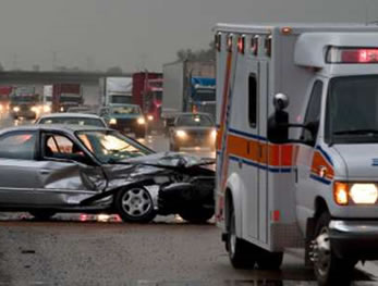 A photo of a car accident scene at the side of the highway. An emergency vehicle sits in front of a heavily damaged passenger car. Highway traffic is seen backed up behind the accident.