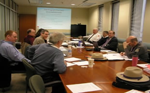 A photo of men and women attending a bi-monthly TIM Steering Committee meeting. Participants are seated at a table in a conference room.