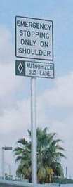 Photo of a post-mounted regulatory sign in the clear zone stating the shoulder is for “Emergency Stopping Only on Shoulder” in black text on a white background. A placard of the same style is mounted below and features a white diamond (corresponding with the pavement marking [not pictured]) and the text “Authorized Bus Lane.”