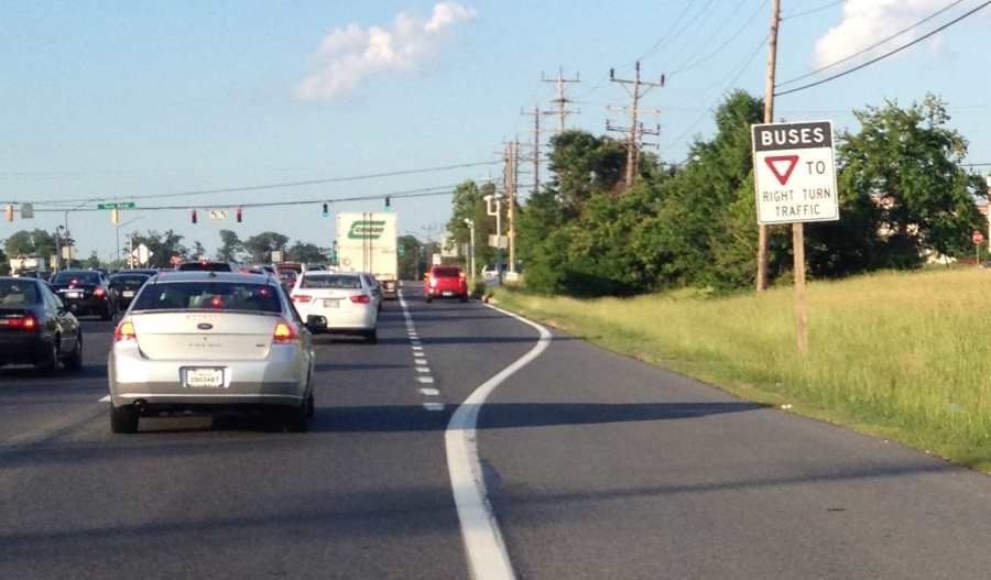 Photo of roadside, post-mounted regulatory sign placed at the beginning of taper for an exclusive right-turn lane at an arterial intersection. The top of the sign states “Buses” in white text on a black background, followed by the image of a yield sign and the text “To Right Turn Traffic” in black text on a white background.