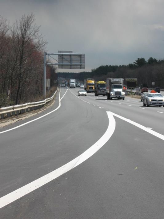 Photo of off-ramp pavement markings in Massachusetts looking upstream from the physical gore. The inside edge of the shoulder lane is solid until the beginning of the taper for the exit lane, where it transitions to a dashed line. The inside edge pavement marking of the shoulder lane then transitions to a dashed line upstream of the physical gore. The outside edge line is solid white and follows the exit lane onto the off-ramp.