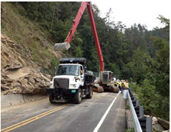 Maintenance crews use bucket loaders and dump trucks to clear a landslide that blocks a roadway.