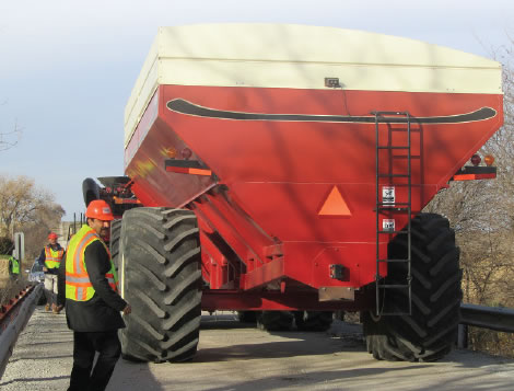 Image of large red and white vehicle with monster truck sized tires on a bridge.  With workman before and after the vehicle.