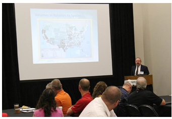 A slides being shown on a screen in a meeting room filled with people.