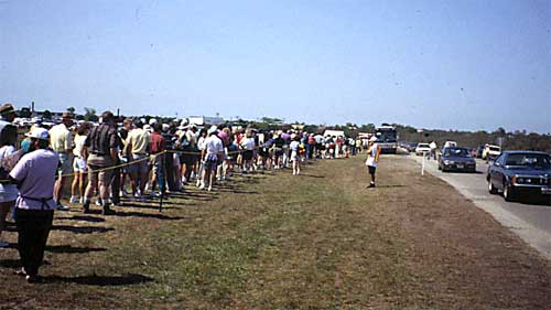 a group of event patrons waiting in line to board a shuttle bus parked on the shoulder of a parking area access road with field personnel present to monitor the patrons