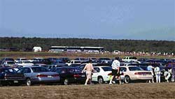 photo showing numerous parked cars, and pedestrians walking along the perimeter of the parking area toward three shuttle buses