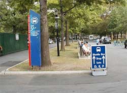 a photo showing several people seated on a bench, located in the median of a local street, near a temporary sign stating "shuttle bus stop – lots D, E, F"