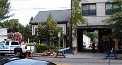 photo showing two police officers standing adjacent to a driveway flanked by a Type 1 barricade on both sides of the driveway.  A temporary disabled parking sign exists in the center of the road and adjacent to the driveway