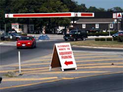 photo showing a police cruiser and Type 1 barricades blocking one roundabout approach, and a guide sign, located in a roundabout gore area, stating "alternate route to aquarium parking"