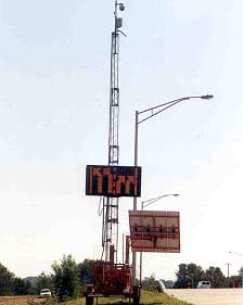 photo showing a device located on the shoulder of a road containing a changeable message sign, displaying two right-turn arrows and two left-turn arrows, an antenna, a closed-circuit television camera at the top of the antenna, and a solar panel