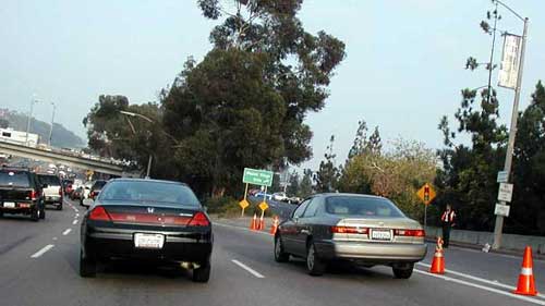 photo showing automobiles in three lanes of divided highway. Orange cones in the gore between the right lane and an exit ramp block the exit. An official wearing an orange safety vest is shown standing on the exit ramp shoulder observing traffic