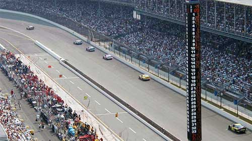 photo of oval raceway, with automobiles racing and spectators seated in the infield and in two tiers of banked seating