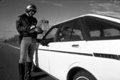 Photograph:    Police officier standing near the driver's side door of an automobile.  The Police officer is writting a citation.    