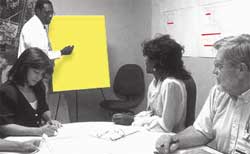 Photograph - Small business meeting.  One individual is about to write on a pad of paper while others in the room are either taking notes or watching the aforementioned individual.