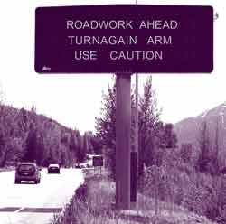 Two cars travel on a highway. A roadside sign warns of imminent roadwork.