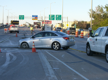 A car on a highway whose front wheel has gone through the surface of the road up to the axle.