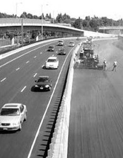 Crews work without interruption on I-84 (Banfield 
Freeway), physically separated from traffic. The picture shows a line of barricades physically separating the workers on the left from the three lanes of oncoming traffic on the right. In the middle of the right hand side of the picture, a pavement grader applies a treatment to the road attended by half a dozen workers.