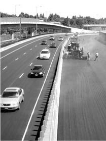 Figure 2 - Crews work without interruption on I-84 (Banfield Freeway), physically separated from traffic. The picture exhibits a view of a construction site along a stretch of the Interstate 84 Banfield Freeway in Portland, Oregon. The picture shows a line of barricades physically separating the workers on the left from the three lanes of oncoming traffic on the right. In the middle of the right hand side of the picture, a pavement grader applies a treatment to the road attended by half a dozen workers.