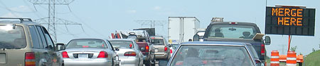 Photo of two lanes of traffic with a Merge Here changeable message sign to the right of the roadway.