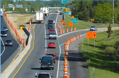 Aerial photo of a divided highway overpass undergoing road construction work. The edge of the right lane and the upcoming exit ramp are bordered by construction barels and the left lanes are defined by jersey barriers.