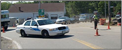 Photo of a local police cruiser blocking one lane of a roadway next to a police officer wearing a yellow-green safety vest behind two orange safety cones blocking a second lane.