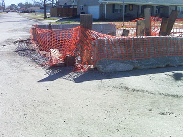 Photo of orange netting wrapped around horizontal concrete blocks and vertical planks on a roadway.