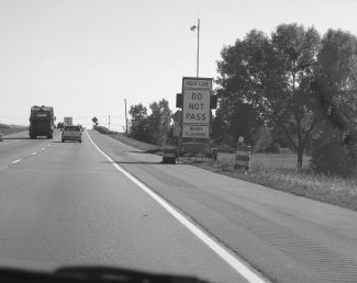 The figure shows a typical trailer setup for the Dynamic Lane Merge System. The trailer is located on the right-hand side of a six-lane Interstate. The trailer has three signs that read Right Lane - Do Not Pass - When Flashing, respectively, from top to bottom. There are two drums with flashing lights in front of the trailer, and a sensor is located approximately 20 feet above the pavement on the top of the trailer.