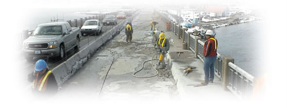 Workmen using jackhammers on a closed lane on a bridge with traffic moving by in a single lane beside them.